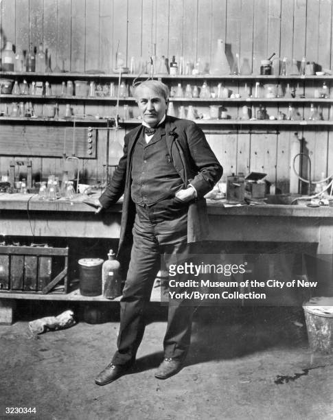 Full-length portrait of American inventor Thomas Alva Edison standing in his laboratory, Menlo Park, New Jersey. Beakers and jars line the shelves.