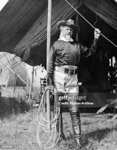 Full-length portrait of American frontiersman and showman William 'Buffalo Bill' Cody holding a rope in front of a tent. He wears a cowboy hat, a...