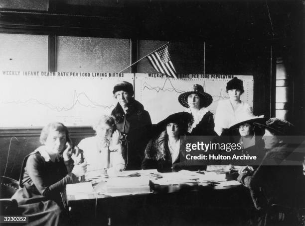 Women sit and stand at a table while enrolling nurses during an influenza epidemic. Graphs post on the wall show mortality rates.