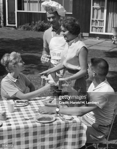 Two couples have a backyard barbecue. A woman serves glasses of beer to her guests in a metal holder while her husband stands behind her, wearing a...