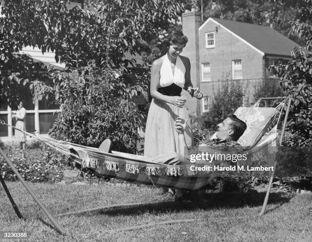 Man lays outdoors in a hammock in his backyard as his wife pours him a glass of beer from a bottle.