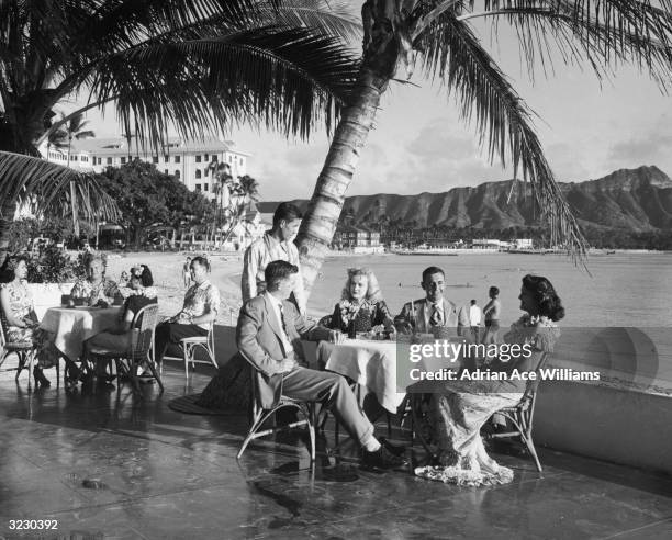 Couples sit outdoors at patio tables, having a meal at a beach side restaurant while a waiter looks on, Waikiki Beach, Honolulu, Hawaii. The women...