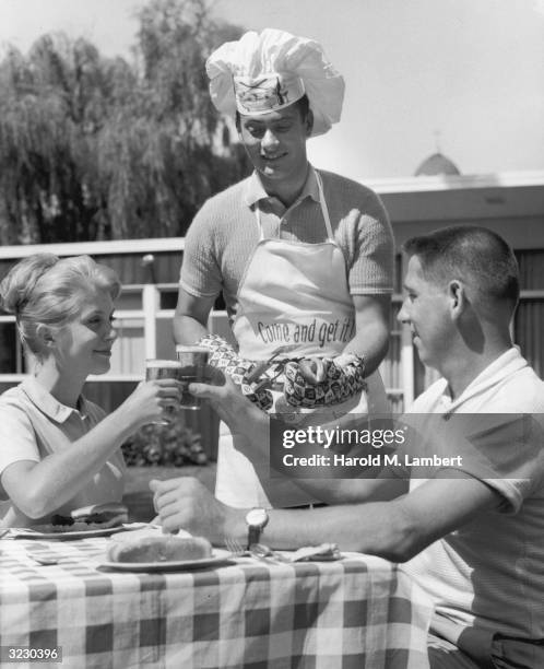 Man in a chef's hat and apron serves hot dogs to a couple sitting at a picnic table, who toast their host with beer during a backyard barbecue.