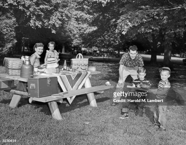 Father crouches behind his sons while they play baseball at a family picnic. One son holds a bat while the other plays catcher with a glove. The...