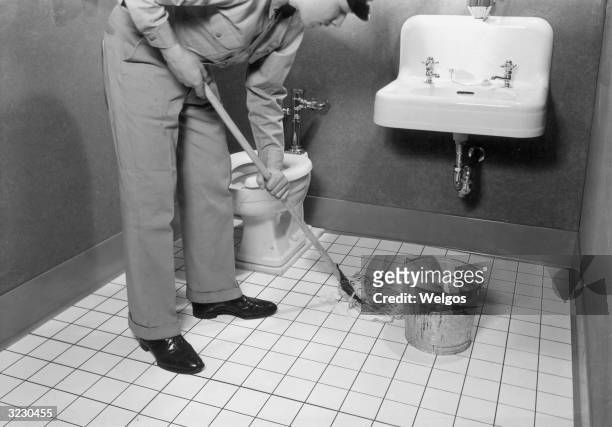 Man wearing a cap and uniform bends down as he mops the floor of a bathroom at a service station. There is a metal bucket full of soapy water on the...