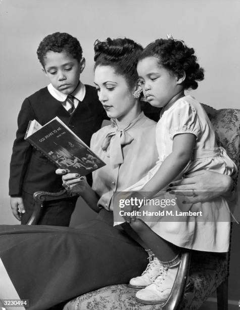 An African-American mother sits in an armchair with her son and daughter, reading a book entitled, 'God Speaks to Me'. The book cover features a...