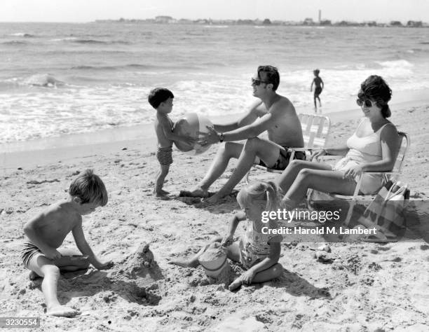 Husband and wife sit on lawn chairs on a beach, watching their sons play in the sand. One of the boys hands a beach ball to his father. The other...