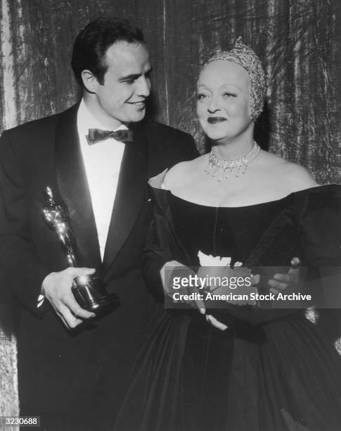 American actors Marlon Brando and Bette Davis smile together backstage at the Academy Awards, Pantages Theatre, Hollywood, California. Brando holds...