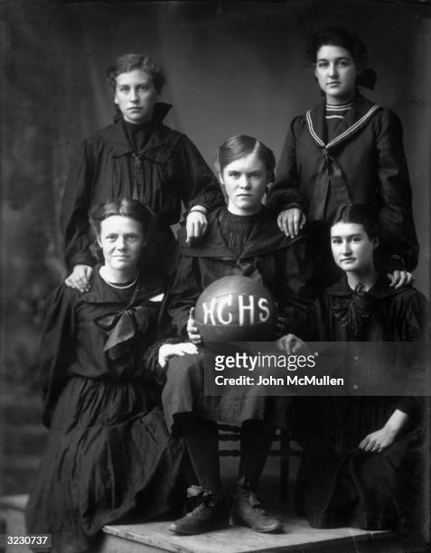 Group studio portrait of five girls on the Harlan County High School women's basketball team, wearing dark dresses and holding a basketball with HCHS...