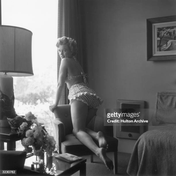 Portrait of American actor Marilyn Monroe , wearing a polka dot two-piece swimsuit, leaning on a chair next to a window in a living room, mid-1950s.