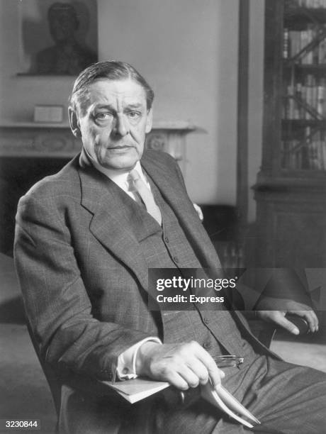 Portrait of American-born poet TS Eliot sitting with a book and reading eyeglasses, around the time of his seventieth birthday.