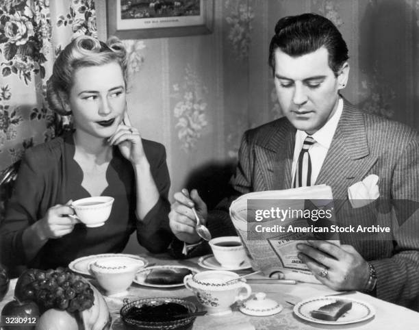 Man sits at a breakfast table reading the newspaper and drinking coffee, as his wife sits next to him and grimaces.