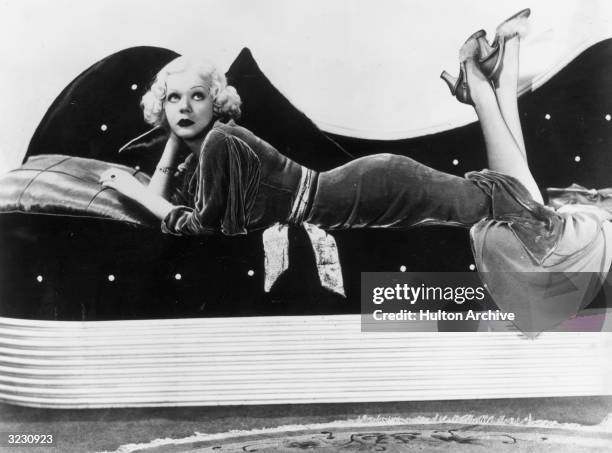 American actor and singer Alice Faye lying on her stomach on a sofa. She wears a velvet dress and high heels trimmed with marabou.