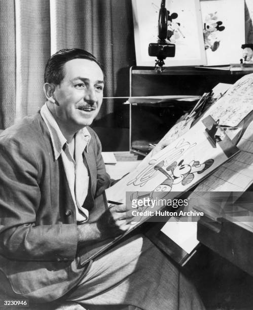 American animator and motion picture studio founder Walt Disney sits at his drawing board in his studio, drawing a sketch of his character Mickey...