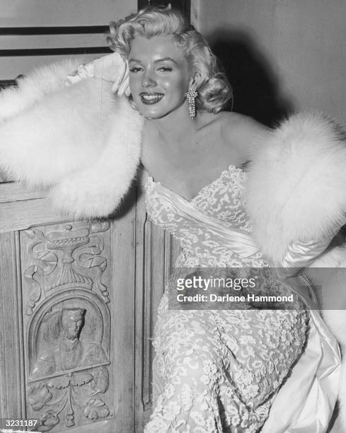 American actor Marilyn Monroe , wearing white fur wrap and strapless evening gown, leans on a wooden cabinet during a party for the film, 'How To...