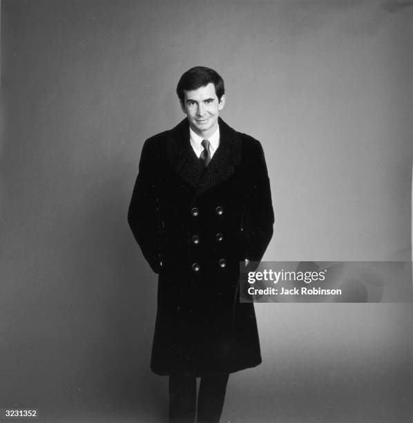 Studio portrait of American actor Anthony Perkins posing in a double-breasted Persian lamb overcoat for Vogue magazine.