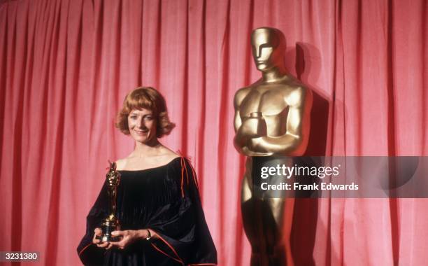 British actor Vanessa Redgrave stands in front of a red curtain and a large Oscar statue, smiling and holding the Oscar she won for Best Supporting...