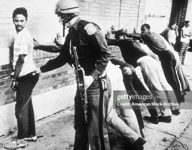 Police frisk an African-American man as other suspects lean against wall on 12th Street, where looting was in progress, during the Detroit riots,...