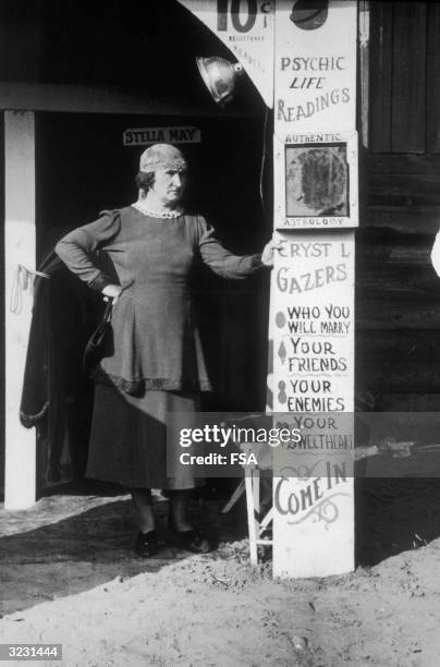 Woman stands with one hand on her hip, and the other leaning on a sign advertising 'PSYCHIC LIFE READINGS...' , with a 'STELLA MAY' sign on a door in...