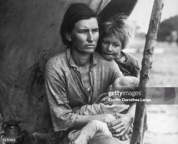 The mother of a migrant family holds a baby while a young girl stands behind her and rests her chin on her shoulder under a lean-to, Nipomo,...