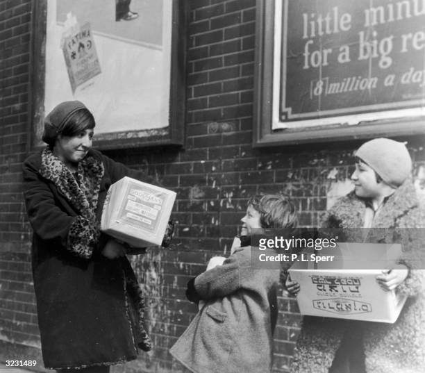 Young Jewish woman and two girls stand outdoors smiling at each other, carrying boxes of matzoth for Passover, Columbia Street, New York City.