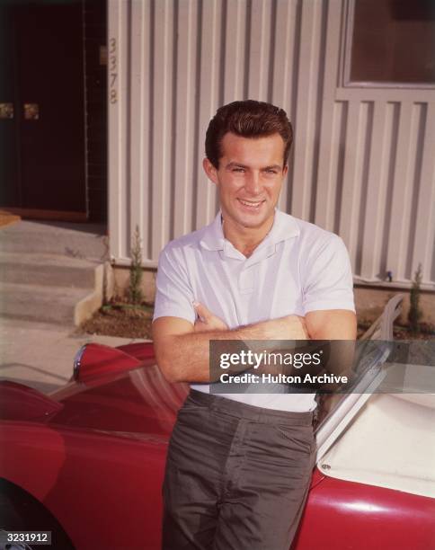 American actor Robert Conrad leans against a red convertible, his arms folded. He wears a white polo shirt.