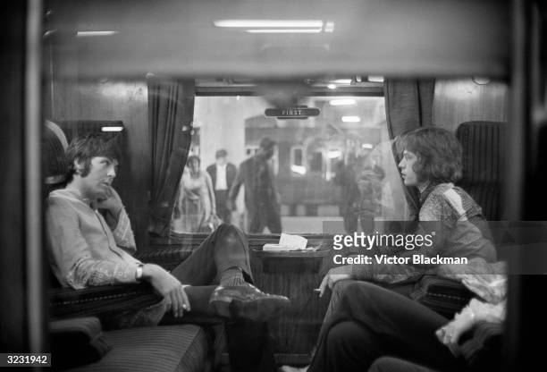 Paul McCartney of the Beatles and Mick Jagger of the Rolling Stones sit opposite each other on a train at Euston Station, waiting for departure to...