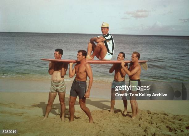 American basketball player Bob Cousy sitting on top of a surfboard carried by four fellow sportsmen in swimming trunks at the beach, circa 1965. The...