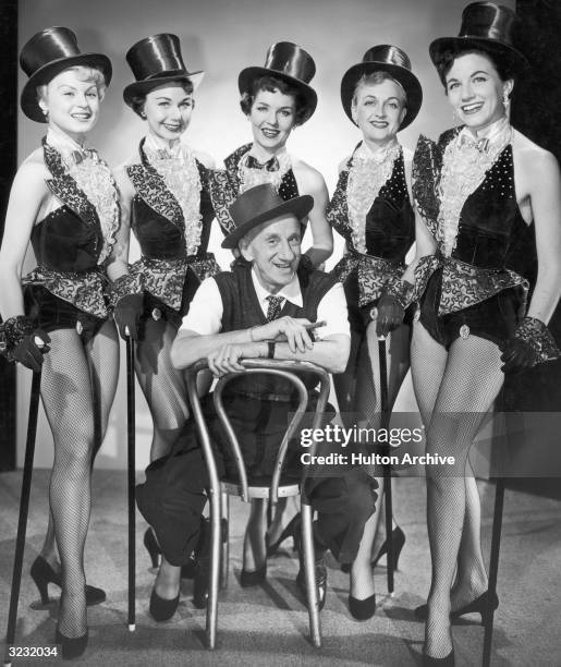 Full-length portrait of American actor and comedian Jimmy Durante sitting backwards in a chair while surrounded by five showgirls wearing identical...
