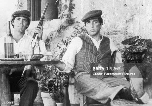 American actor Al Pacino sits with Italian actor Franco Citti holding a rifle at an outdoor table in director Francis Ford Coppola's film, 'The...