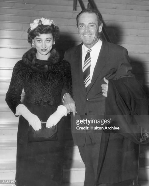 American actor Henry Fonda smiles with his fourth wife, Afdera Franchetti. She wears white gloves and holds her purse. He holds his coat over one arm.