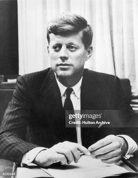 Portrait of US senator John F Kennedy of Massachusetts sitting at his desk with a pen and paperwork.