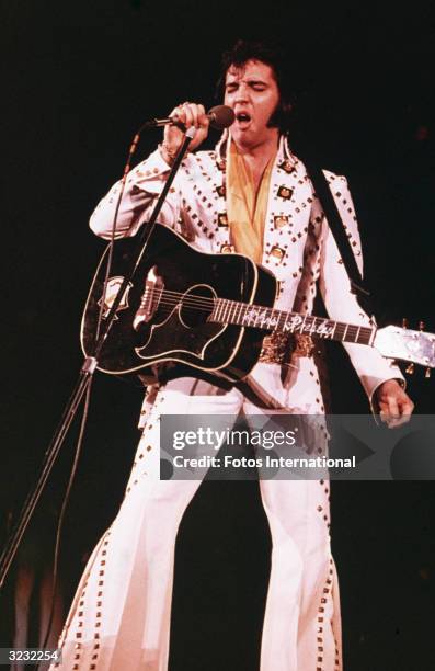 American rock singer Elvis Presley , wearing a white rhinestone-studded suit and strapped guitar, singing into a microphone with his eyes closed.