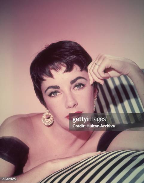 Promotional headshot portrait of British-born actor Elizabeth Taylor leaning on a striped sofa in a sleeveless gown and short hair.