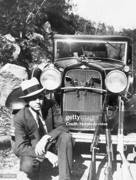 Portrait of American outlaw Clyde Barrow crouching in front of a car with a handgun hanging from the hood ornament, two handguns in the front grill...
