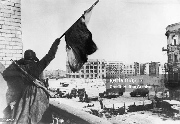 Russian soldier waves a flag while standing on a balcony overlooking a square, where military trucks gather, during the Battle of Stalingrad, World...