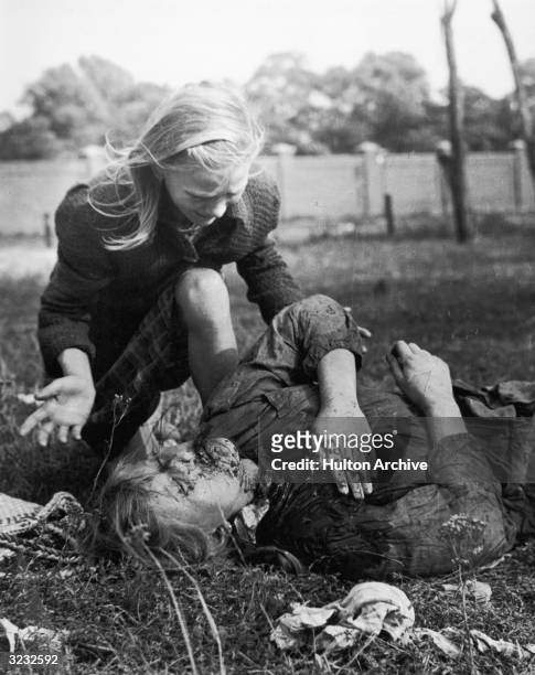 Young girl cries over her wounded and bleeding sister, who lies on the ground with her hand covered over a chest wound, during the Nazi invasion of...