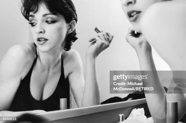 Portrait of future American pop singer Madonna looking away from her reflection while sitting in front of a mirror with a tube of lipstick in her...