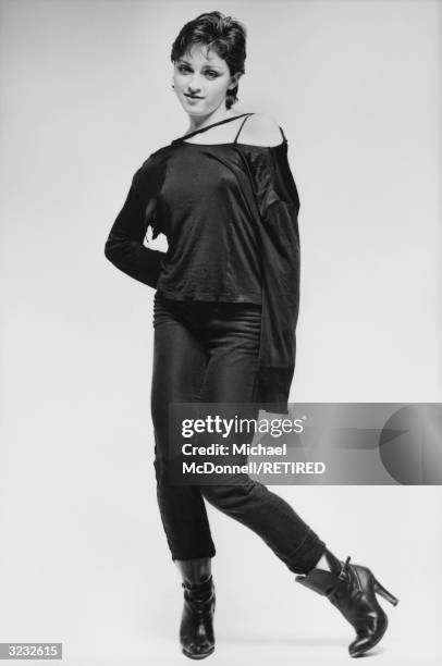 Full-length studio portrait of future American pop singer Madonna standing knock-kneed in a ripped, black off-the-shoulder shirt, tight black Levi's...