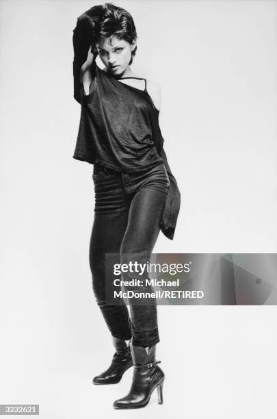 Full-length studio portrait of future American pop singer Madonna standing with her hand behind her head and wearing a ripped, black,...