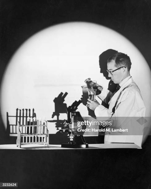 Studio image of a scientist standing in a spotlight beside a microscope and a test tube holder, pouring a solution from one beaker to another. He...