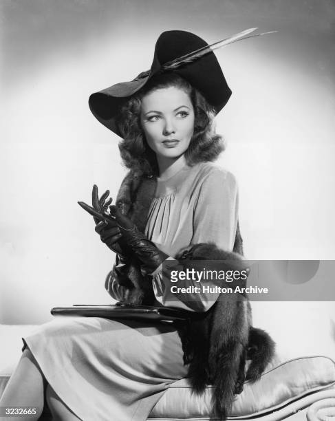 Studio portrait of American actor Gene Tierney seated on a divan. She is wearing a hat with a feather, a mink stole, and leather gloves.