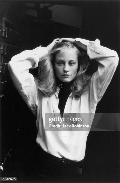 Portrait of American actor and model Cybill Shepherd posing in a doorway with her hands behind her head, New York City. She wears a white blouse.