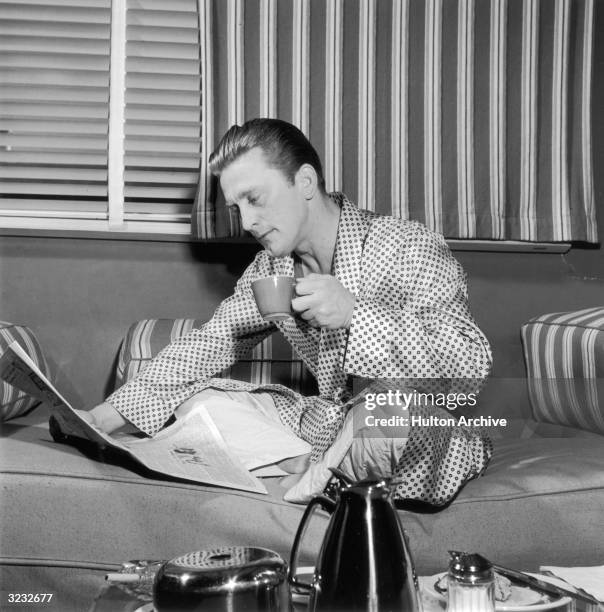 American actor Kirk Douglas, wearing a silk robe and pajamas, sits cross-legged on a sofa as he reads a newspaper and drinks coffee. A room service...