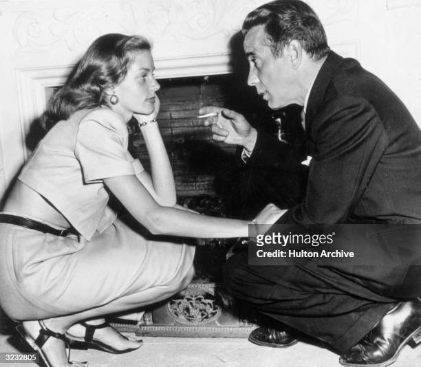 Married American actors Lauren Bacall and Humphrey Bogart kneel in front of a fireplace in their home, California. Bogart is holding a cigarette, and...
