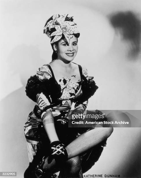 Studio portrait of American dancer and choreographer Katherine Dunham smiling and sitting with her legs crossed. Dunham is wearing a dance costume...