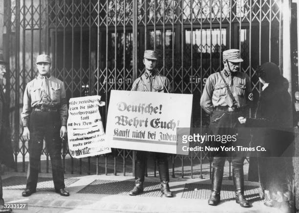 Nazi troops hold anti-Semitic placards, in front of a locked storefront in the organised boycott of German Jewish businesses, Berlin, Germany. One of...