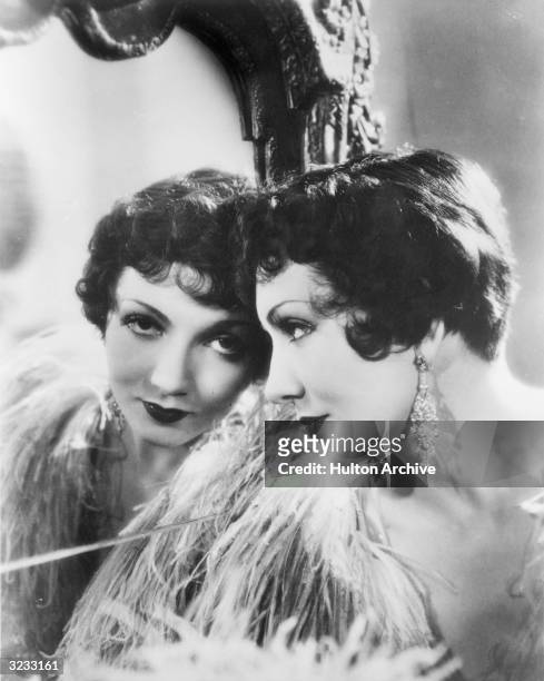 Headshot portrait of American actor Claudette Colbert looking into a mirror. She is wearing a feather boa and dangling earrings.