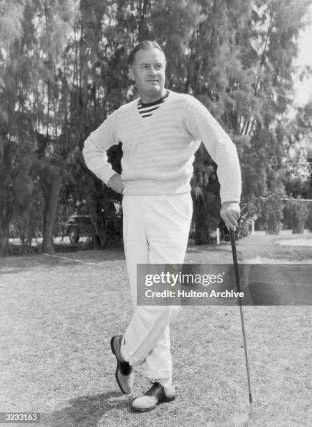 Full-length image of British-born entertainer Bob Hope leaning on a golf club outdoors. He is wearing white pants, a striped shirt, and a v-neck...