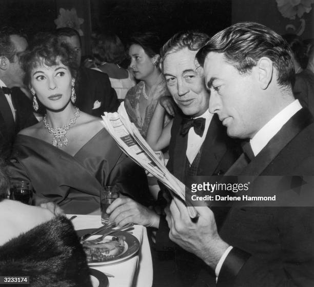 American actor Gregory Peck reads a newspaper review of director John Huston's film, 'Moby Dick' while his wife, Veronique, and Huston himself look...